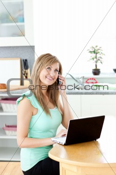 Smiling woman on phone using a laptop in the kitchen