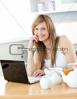 Smiling woman having breakfast and using her laptop looking at t