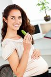 Bright pregnant woman eating a gherkin in her office