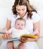 Happy mother showing a book to her baby sitting in the sofa at home