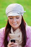 Pretty young woman wearing cap and scarf sending a message with her cellphone on the grass in a park