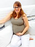 Smiling beautiful pregnant woman sitting on sofa and holding her belly.
