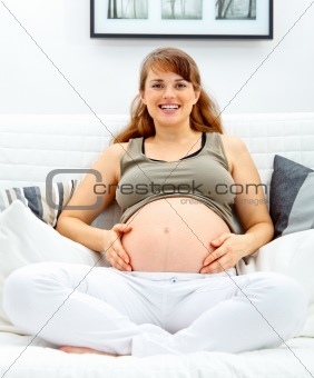 Smiling beautiful pregnant woman sitting on sofa and touching her belly.
