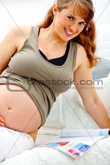 Smiling beautiful pregnant woman sitting on sofa with magazine.
