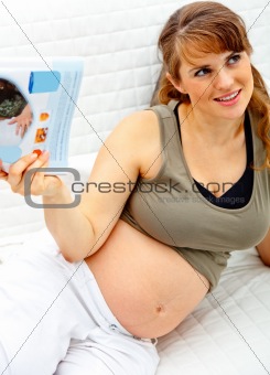 Smiling beautiful pregnant woman relaxing on couch with magazine.
