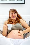 Laughing beautiful pregnant woman sitting on sofa and  holding cup of tea in hand
