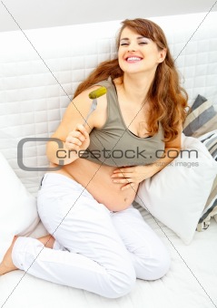 Smiling  beautiful pregnant female sitting on divan and eating pickle

