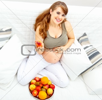Smiling  beautiful pregnant female sitting  on sofa and  holding fruit in hand

