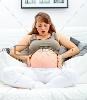 Surprised  beautiful pregnant woman sitting on sofa and holding her belly.
