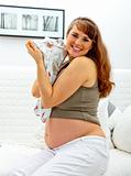 Smiling beautiful pregnant woman sitting on sofa with baby clothes  in hands.
