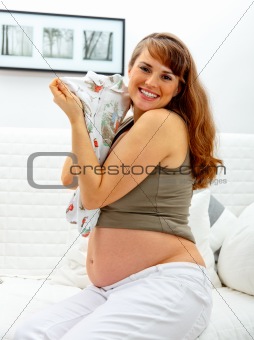 Smiling beautiful pregnant woman sitting on sofa with baby clothes  in hands.
