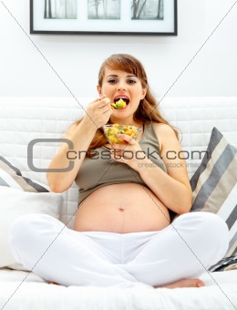 Happy beautiful pregnant woman sitting on sofa and eating fruit salad
