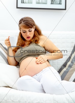 Surprised beautiful pregnant woman sitting on sofa and  holding cup of tea in hand
