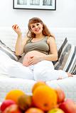 Smiling  beautiful pregnant woman sitting on divan and  holding fruit in hand
