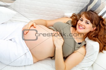 Smiling beautiful pregnant female relaxing on sofa and  holding her belly.
