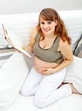 Smiling beautiful pregnant woman sitting on sofa with book.
