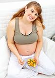 Happy beautiful pregnant woman sitting on sofa with fruit salad  in hand.
