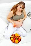 Smiling  beautiful pregnant female sitting  on couch and  holding fruit in hand
