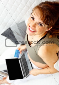 Smiling beautiful pregnant female on sofa with the laptop and  credit card.

