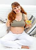 Smiling beautiful pregnant female sitting on sofa with fruit salad  in hand
