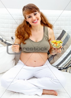 Smiling beautiful pregnant female sitting on sofa with fruit salad  in hand

