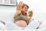 Embarrassed beautiful pregnant woman sitting on sofa and  holding jar of pickles in hands
