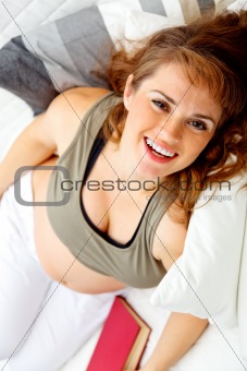 Smiling beautiful pregnant female relaxing on sofa with book.
