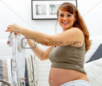 Smiling beautiful pregnant female sitting on sofa with baby clothes  in hands.
