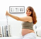 Happy beautiful pregnant woman sitting on sofa with baby clothes  in hands.
