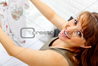 Smiling beautiful pregnant woman sitting on sofa with baby clothes  in hands. Closeup.
