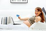 Smiling beautiful pregnant woman using credit card to shop from net and showing thumbs up gesture
