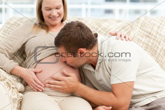 Future dad kissing the belly of his wife sitting on the sofa
