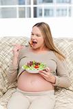 Cute pregnant woman eating a salad sitting on the sofa