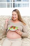 Happy pregnant woman enjoying a salad sitting on the sofa