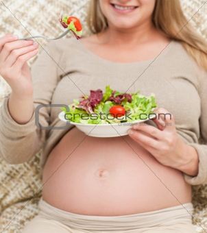 Cute pregnant woman eating a salad sitting on the sofa