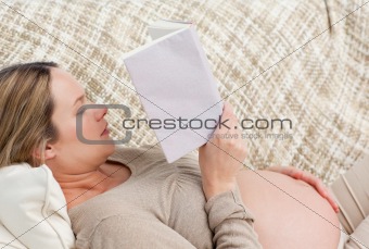 Cute pregnant woman reading a book lying on the couch