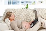 Smiling pregnant woman using her laptop lying on the sofa