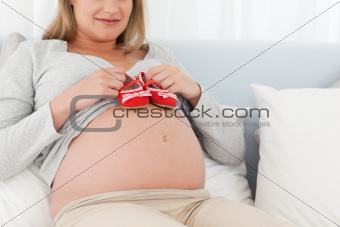Cute future mom putting baby shoes on her belly relaxing on a bed