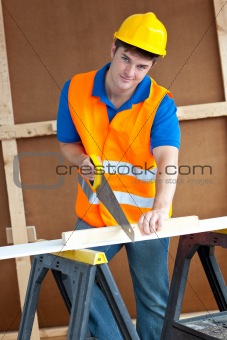 Charismatic male worker wearing a yellow hardhat sawing a wooden