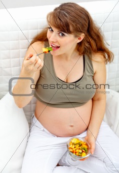 Smiling beautiful pregnant woman sitting on sofa and eating fruit salad
