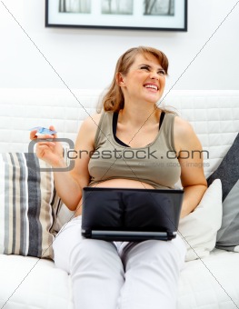 Laughing beautiful pregnant woman sitting on sofa with laptop and credit card
