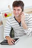 Attractive man talking on phone using his laptop in the kitchen