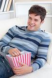 Cheerful young man eating popcorn lying on the sofa