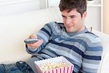 Bright young man eating popcorn and holding a remote lying on th