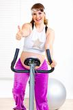 Smiling beautiful pregnant woman working out on static bicycle and showing thumbs up gesture
