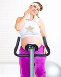 Beautiful pregnant woman wiping her face with towel after training on static bike
