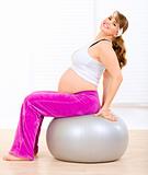 Smiling beautiful pregnant woman doing pilates exercises on gray ball at home
