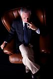 Pensive mature businessman seated on a chair, isolated on black background. Studio shot.