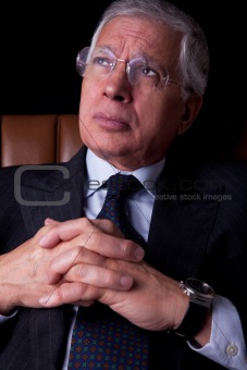 Pensive mature businessman seated on a chair, looking up, isolated on black background. Studio shot.
