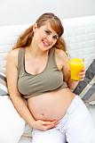 Smiling beautiful pregnant woman sitting on couch with glass of juice  in hand
